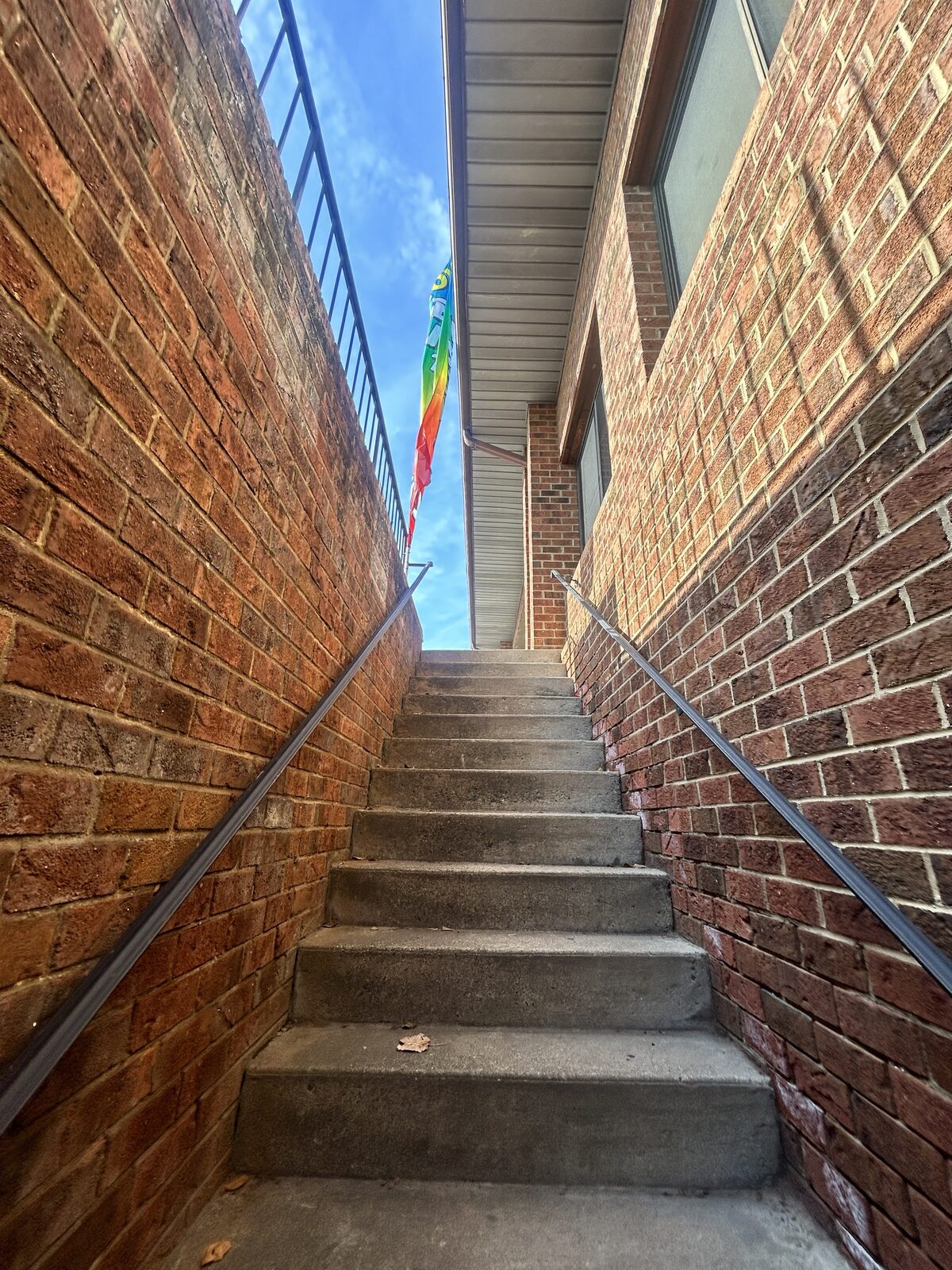 Narrow brick stairwell between two walls, a handrail on each side, seen from the bottom landing looking up.